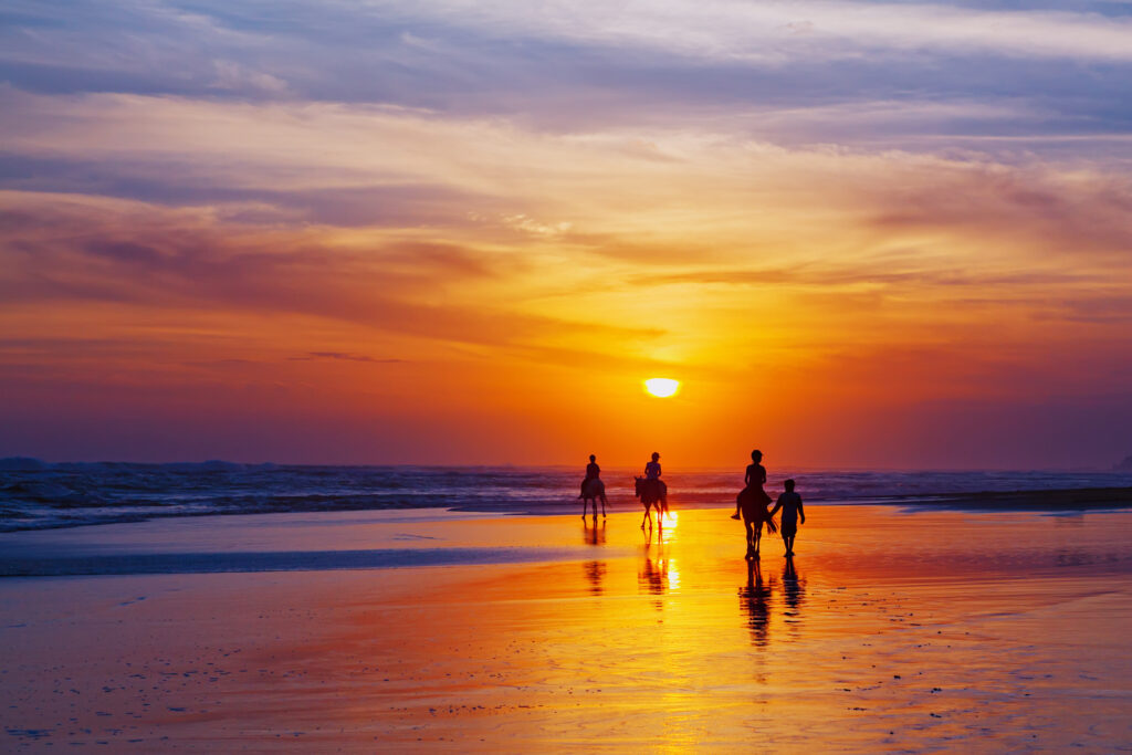Black silhouette of happy family having horse riding adventure on sand sea beach on background of sunset sky. Active parents and people outdoor activity on tropical summer vacations with children.