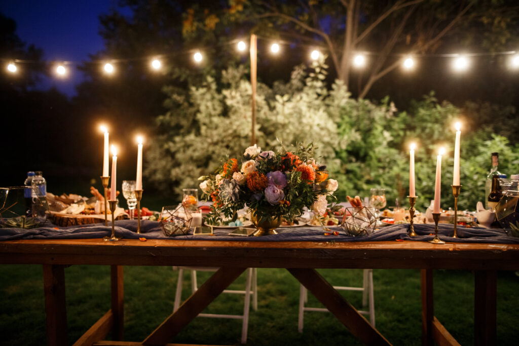 A wooden table with a flower bouquet in the middle and a lots of cutlery and candles alongside it. Garland and some trees are at the background.