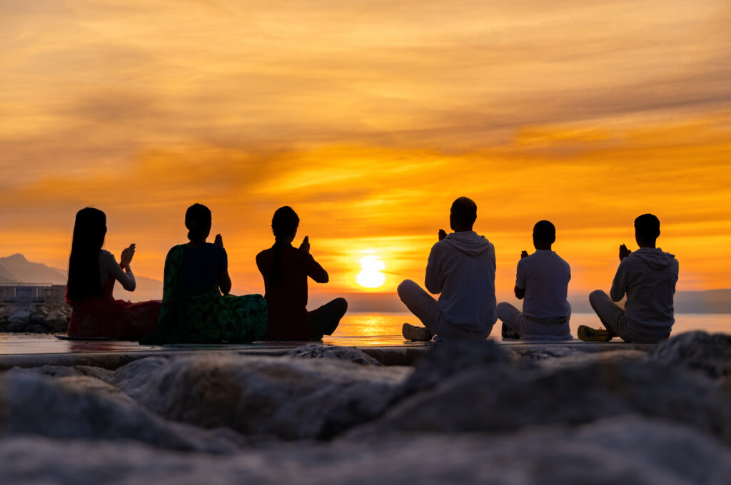 Silhouettes of group of young people meditating on the sunrise sun at the sea. Prayer, meditation and yoga in the nature.
