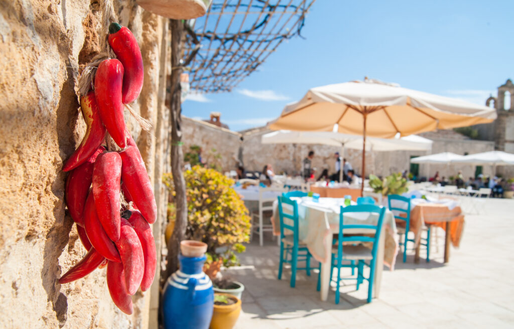 Small pile of fake red peppers hanging on the wall of a typical stone house in the fishing village Marzamemi, Sicily, and the main square with tables of a restaurant